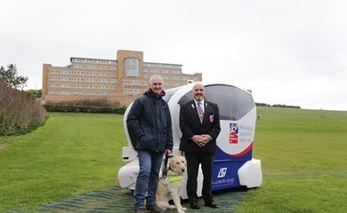Three Men and a guide dog, stand with an electric pod car on grass, with a large building behind them 