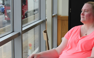 Lady with blonde hair wearing a pink t shirt, sitting in a wheelchair looking out of a window