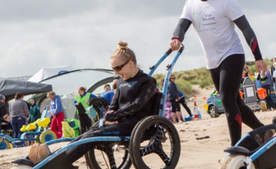 Image shows a women sitting in a sand wheelchair with a man pushing them along. They are both wearing wetsuits. They are on a beach and there are people around in the background.
